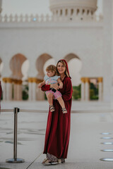Dubai,  Unite dArab Emirates - October 19, 2019 - A smiling woman in a red dress holding a child, standing in front of a mosque's white arcade.