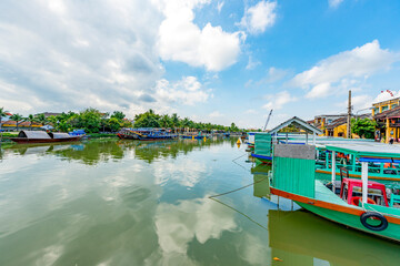Obraz premium Wooden boats on the Thu Bon River in Hoi An Ancient Town (Hoian), Vietnam. Yellow old houses on waterfront reflected in river. Riverside Scenes in the Historic District of Hoi An, Vietnam