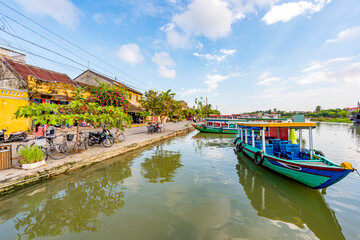 Fototapeta na wymiar Wooden boats on the Thu Bon River in Hoi An Ancient Town (Hoian), Vietnam. Yellow old houses on waterfront reflected in river. Riverside Scenes in the Historic District of Hoi An, Vietnam