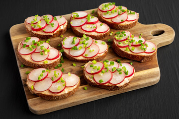 Homemade Lemon Radish Tartine on a wooden board on a black background, side view.