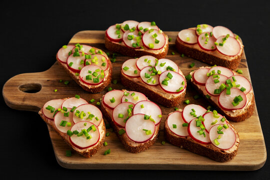 Homemade Lemon Radish Tartine on a wooden board on a black background, side view.