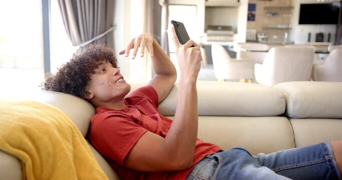 A young biracial man is lounging on a sofa at home, interacting with his smartphone