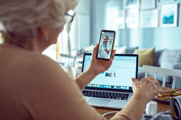 Elderly woman having a video call with her doctor on a smartphone with a laptop open in the background