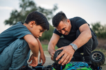 Dad and his boy are fixing a broken remote toy car while crouching outdoors.