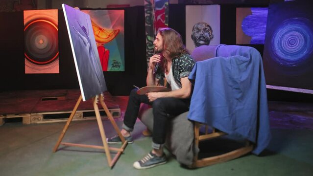 Side view of original painter sitting on wooden rocking chair covered with blue cloth inside art studio. Pensive caucasian artist examining masterpiece and prouding of work indoors.