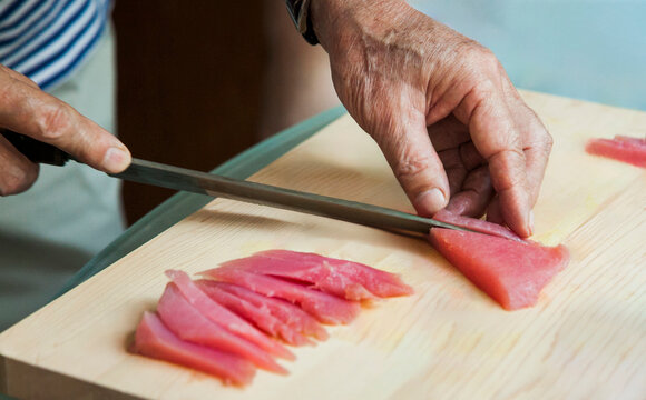 Hands of a senior man, grandfather, indoors, cutting up a piece of red tuna on a wooden cutting board to prepare sashimi - Powered by Adobe