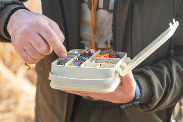 The fisherman's hands are holding a plastic box with a variety of lures and fishing hooks
