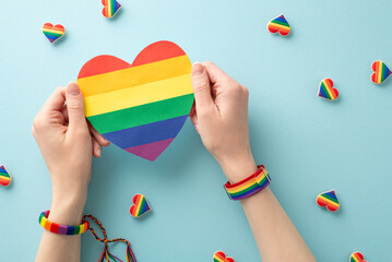 This powerful first person top view image represents the contributions of the LGBT community throughout history, featuring a rainbow heart held by hands in symbolic wristlet, light blue backdrop - obrazy, fototapety, plakaty