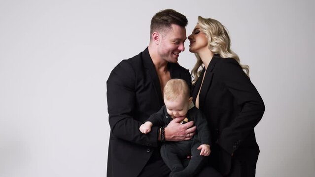 Caucasian couple in love wear black jackets and kissing tenderly. Beautiful portrait of family of three in studio. White backdrop.