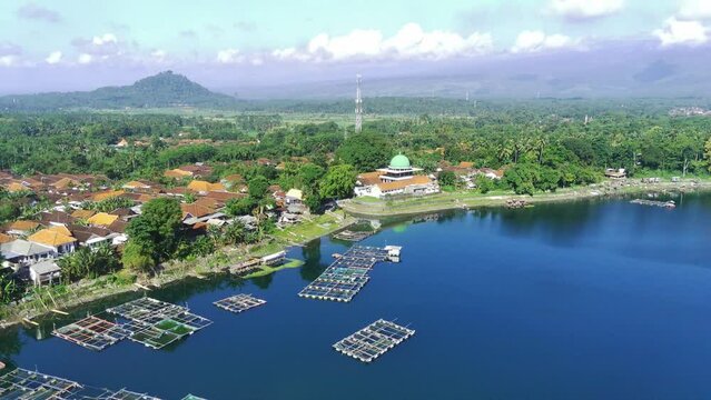 An aerial view of a serene lake with a white mosque with a dome and tall minaret in the background. Lush greenery surrounds the lake. Near Ranu Pakis, Lumajang, East Java, Indonesia.