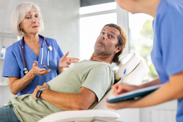 Fototapeta premium Middle-aged man is sitting in chair at clinic reception and carefully answers mature female doctor questions about his well-being, health complaints, nurse records data in client medical record