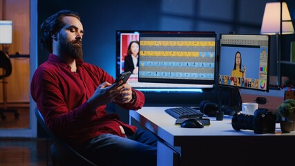 Photographer uses retouching software to edit pictures on computer monitors, taking a break after working too much. Photo editor tired after doing color grading on images, relaxing by using smartphone