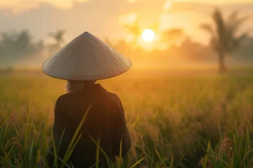 Foto op Plexiglas A contemplative silhouette of a woman in traditional attire against the backdrop of a sunset over a rice field © ChaoticMind