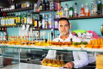 Friendly young male bartender offering appetizing pinchos in comfortable pub