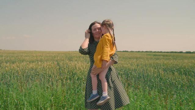 Mom and little daughter are walking in green wheat field, hugging and kissing. A child and mom are walking in wheat field. Happy family travel. Baby is in mom's arms. Farmer woman and child in field