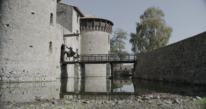 Hooded man on horseback riding into castle as drop gate closes behind him