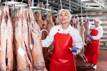 Smiling female butcher showing muttton carcass in meat storage