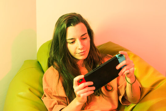 A relaxed gamer is captured in a moment of focus while playing on a portable gaming console, nestled in a cozy, brightly colored chair