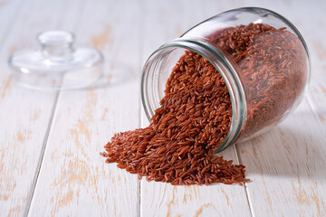 Raw red rice spill out of a glass storage jar on a white wooden table.