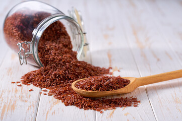 Raw red rice in wooden scoop and in glass storage jar on a white wooden table.