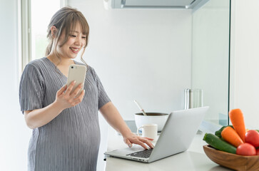 キッチンで料理をしながらパソコンとスマホを使う妊婦の女性
