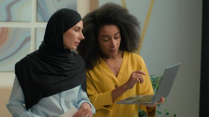 Two multiracial women diverse girls colleagues coworkers businesswomen talk discuss business project on laptop in office African American mentor consult teach Arabian muslim islamic female in hijab
