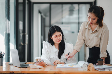 Two female real estate professionals discuss project details with a model house on the table, symbolizing project planning and teamwork.