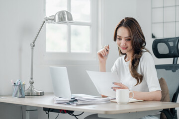 Obraz na płótnie Canvas Young professional woman analyzing paperwork with a focused expression, seated at her home office desk with a laptop and coffee mug.