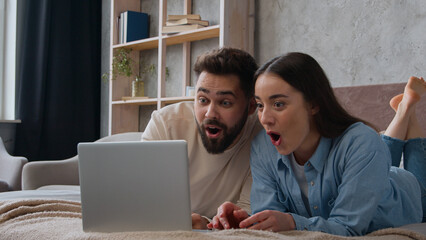Excited Caucasian married couple lying on bed at home win laptop achievement read amazing news...