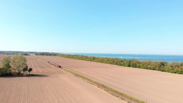 Aerial: steam narrow gauge railway in the countryside  passing by sown fields and the sea in the background