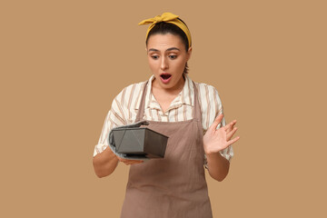 Portrait of shocked young housewife in apron with baking form and mitt on brown background