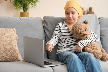 Little girl after chemotherapy with toy bear watching cartoons on laptop at home. International...