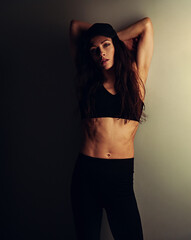 Sport sexy body beautiful slim woman with long hair posing in black sport bra, summer cap showing the shoulders, abs, arms, standing on studio wall background with empty copy space. Lifestyle vintage