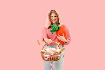 Beautiful young happy woman in bunny ears with carrot-shaped toy and Easter basket on pink...
