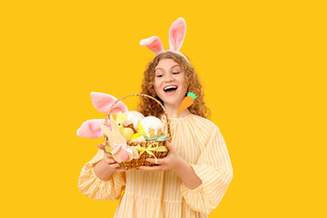 Beautiful young shocked woman in bunny ears with Easter basket on yellow background