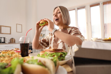 Beautiful woman with tasty burger at table in kitchen. Overeating concept