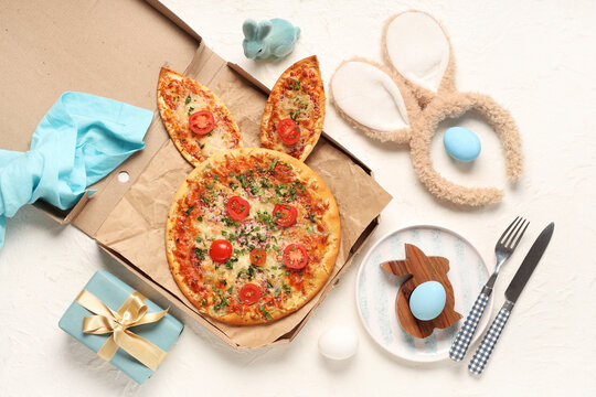 Cardboard box of Easter pizza with bunny ears, painted eggs and gift on white background