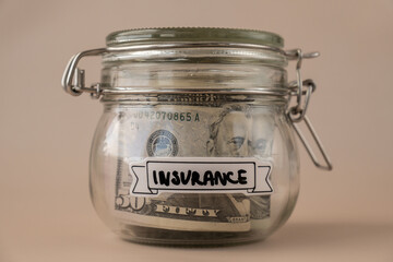 Saving Money In Glass Jar filled with Dollars banknotes. INSURANCE transcription in front of jar. Managing personal finances extra income for future insecurity. Beige background