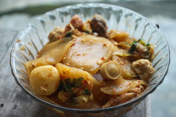 Seblak is a typical Indonesian dish with a savory and spicy taste. cooked with vegetables, sausage, eggs, chicken feet, seafood and processed beef. a bowl of seblak on a wooden table.