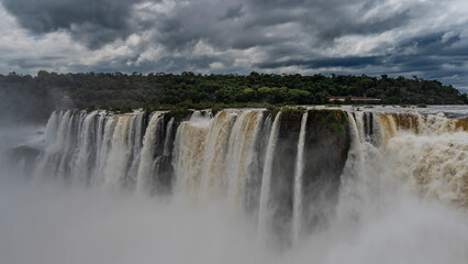 The incredible powerful waterfall - the Devil's Throat. Streams of foaming white water fall into the abyss. Green vegetation on the river bank. Clouds in the sky. Iguazu Falls. Argentina.