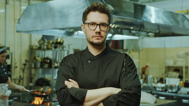 Confident head chef in uniform and glasses standing in the restaurant kitchen and posing at camera with arms crossed. Video portrait
