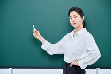 Wearing white heating, a beautiful Asian female teacher is holding a chalk in front of a green chalkboard and giving an Internet lecture with a confident expression and pose.
