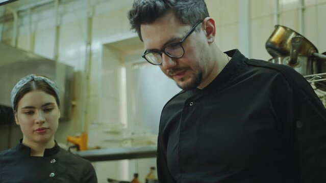 Skilled chef explaining cooking technique and giving culinary class to young female trainee listening attentively and holding copybook during internship in restaurant kitchen