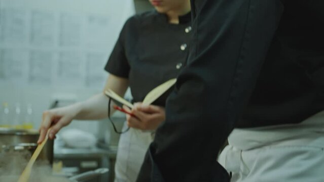 Master chef mincing garlic, telling recipe and showing cooking steps to young female trainee taking notes in copybook as guiding her during internship in restaurant kitchen