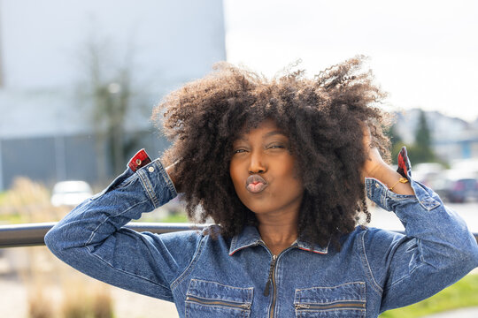 Captivating outdoor shot of a joyful African American woman wearing a denim jacket, making a playful kissy face. She stands confidently with hands in her hair, enhancing her beautiful, textured afro