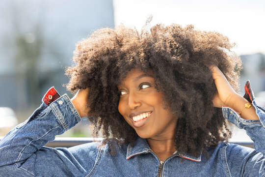 An exuberant African American woman with a radiant smile and a stunning afro hairstyle is caught in a candid moment. She playfully touches her hair while clad in a classic denim jacket, her expression