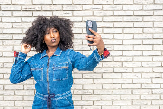 This vibrant image captures a young African American woman in a trendy denim outfit taking a selfie. Her playful pout and voluminous afro hairstyle stand out against the textured white brick wall