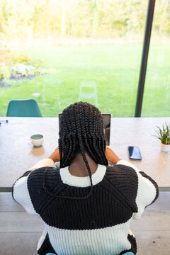 A serene scene featuring the back view of a black woman with braided hair working attentively on her tablet in a bright and airy home office. The space overlooks a lush garden, combining productivity