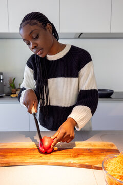 A woman with braided hair is immersed in cooking, meticulously slicing a tomato on a wooden cutting board. The setting is a contemporary kitchen, where the simple act of food preparation becomes an