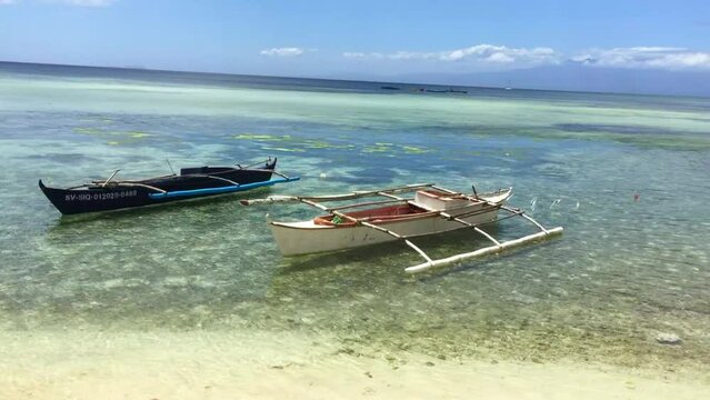 Two traditional Filipino boats are anchored on the Siquijor Island coastline. The wooden boats are in the crystal clear water. The blue sky with some small clouds. A sunny day on the beach.